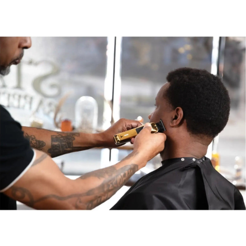 A barber with tattooed arms uses an electric clipper to trim a client's hair in a barbershop setting.