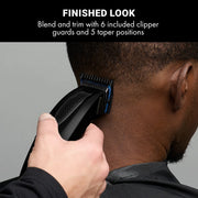A close-up of a person receiving a haircut with a black electric clipper against a gray background, with a black text banner at the top reading 'FINISHED LOOK Blend and trim with 6 included clipper guards and 5 taper positions'.