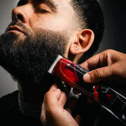 A close-up of a man receiving a beard trim, with a barber's hand holding a red and black electric clipper against his beard. The photo is framed within a white border on a white background.