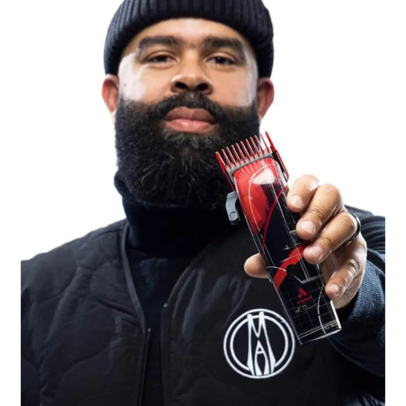 A man with a full beard and black beanie holds up a clear plastic clipper guide comb with red and black blades against a plain white background.